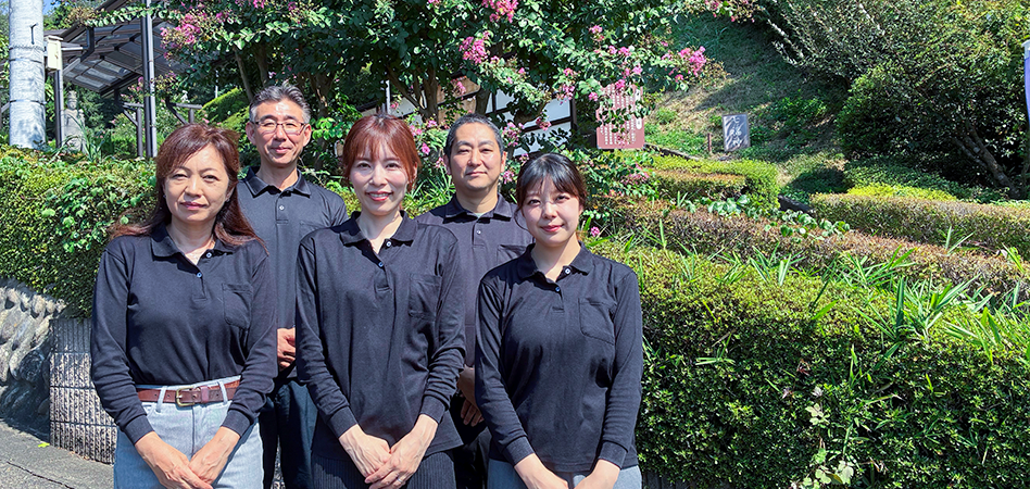 ペット霊園のスタッフ集合写真。緑に囲まれたペット霊園で、制服を着たスタッフ5名が笑顔で並んで撮影されている。